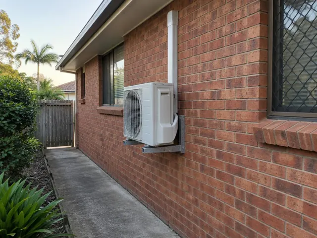 Wall-mounted air conditioner outdoor unit on a brick house exterior beside a side path in Helensvale